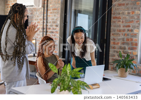 Diverse female coworkers clapping and laughing around laptop and smartphone on modern office desk Diverse female coworkers clapping and laughing around laptop and smartphone on modern office desk 140563433