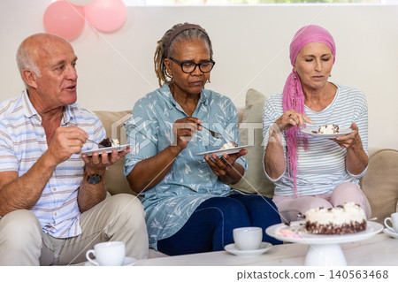 Diverse senior friends sitting on sofa at home eating cake and holding cups under balloons Diverse senior friends sitting on sofa at home eating cake and holding cups under balloons 140563468