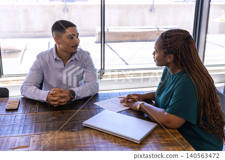 Diverse coworkers discussing notes with document at office conference table with laptop smartphone Diverse coworkers discussing notes with document at office conference table with laptop smartphone 140563472