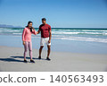 Diverse couple standing on sandy shore by gentle surf in pink jacket and athletic shoes 140563493