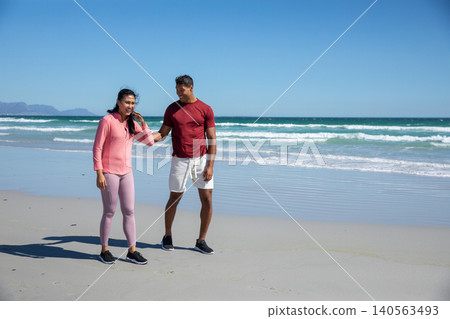 Diverse couple standing on sandy shore by gentle surf in pink jacket and athletic shoes Diverse couple standing on sandy shore by gentle surf in pink jacket and athletic shoes 140563493