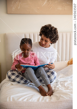 African american mother with daughter reading blue book on duvet-covered bed with teddy bear African american mother with daughter reading blue book on duvet-covered bed with teddy bear 140563507