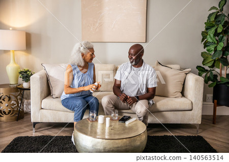 Diverse couple discussing pill bottles and water beside smartphone on beige sofa in living room Diverse couple discussing pill bottles and water beside smartphone on beige sofa in living room 140563514