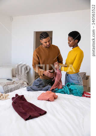 Hispanic and African American couple folding colorful clothes on bed in bedroom with cardboard box Hispanic and African American couple folding colorful clothes on bed in bedroom with cardboard box 140563522