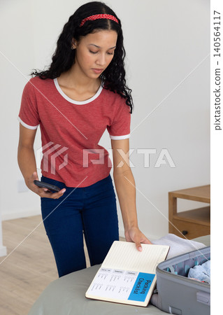 Teenage girl with smartphone and leaning over packing checklist by open suitcase in bedroom Teenage girl with smartphone and leaning over packing checklist by open suitcase in bedroom 140564117