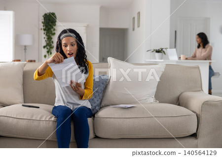 Mother and teenage daughter on sofa, typing on laptop holding mug and daughter opening envelope Mother and teenage daughter on sofa, typing on laptop holding mug and daughter opening envelope 140564129