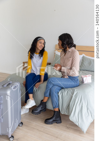 Diverse mom and daughter sitting on bed near silver suitcase, pastel pink gift box 140564130