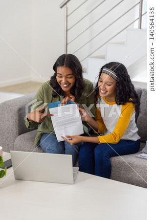 Diverse mom and teenage daughter sitting on sofa in living room examining document Diverse mom and teenage daughter sitting on sofa in living room examining document 140564138
