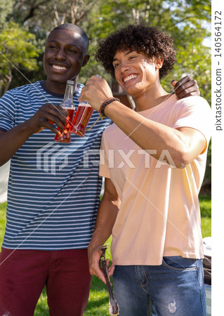 Diverse male friends toasting with glass bottles of drink in backyard near picnic table 140564172