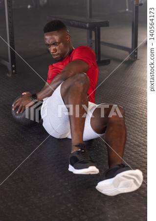 African American man twisting with medicine ball on gym floor in red shirt and gray shorts African American man twisting with medicine ball on gym floor in red shirt and gray shorts 140564173