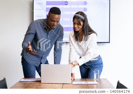 Diverse coworkers leaning over table in boardroom analyzing data on laptop, screen with charts Diverse coworkers leaning over table in boardroom analyzing data on laptop, screen with charts 140564174