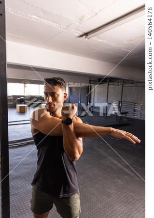 Man stretching arm across chest inside fitness center near battle ropes and weight plates Man stretching arm across chest inside fitness center near battle ropes and weight plates 140564178