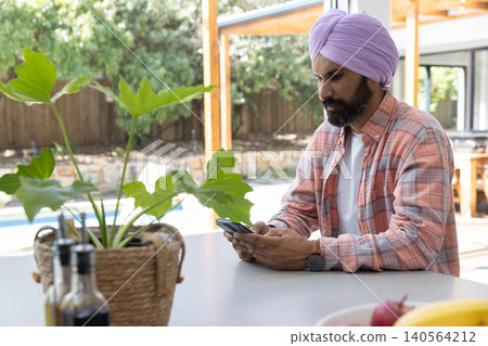 Indian man wearing lavender turban, plaid shirt using smartphone at patio counter, copy space Indian man wearing lavender turban, plaid shirt using smartphone at patio counter, copy space 140564212