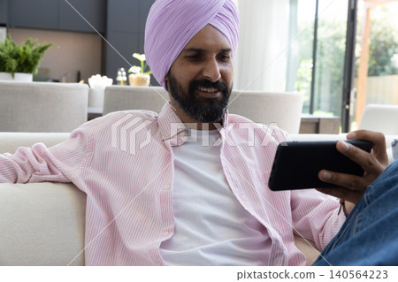 Indian man sitting on sofa in living room holding tablet and wearing turban Indian man sitting on sofa in living room holding tablet and wearing turban 140564223