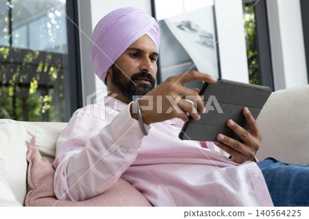 Mid adult indian man reclining on sofa with cushion at home holding tablet next to window Mid adult indian man reclining on sofa with cushion at home holding tablet next to window 140564225