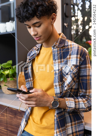 Man in plaid shirt standing at kitchen checking smartphone beside ceramic mugs and plant 140564232