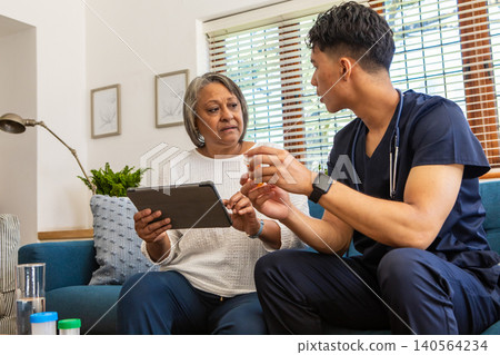 Nurse wearing scrubs, sitting at home with african american senior holding tablet, pill bottle Nurse wearing scrubs, sitting at home with african american senior holding tablet, pill bottle 140564234