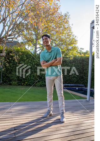 Man in early twenties standing with folded arms on wooden deck near hedge and pool cover Man in early twenties standing with folded arms on wooden deck near hedge and pool cover 140564242