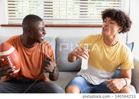Diverse male friends sitting on couch in living room holding football, raising bottled beverage 140564253