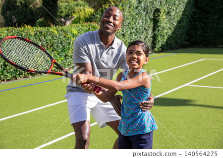 Diverse father and daughter practicing tennis swing on tennis court with red-and-black racket Diverse father and daughter practicing tennis swing on tennis court with red-and-black racket 140564275