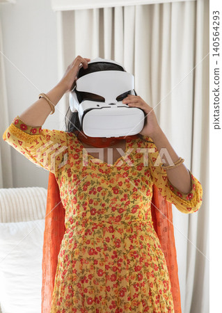 Indian woman adjusting vr headset while standing in bedroom beside bed with pillows Indian woman adjusting vr headset while standing in bedroom beside bed with pillows 140564293