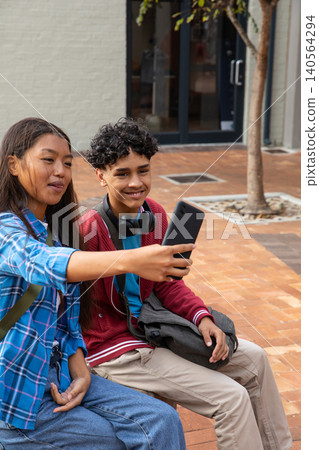 Diverse siblings holding smartphone and taking selfie on paved courtyard bench with backpack 140564294
