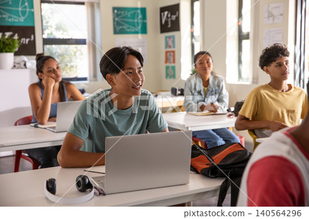 Asian teenage male student studying on laptop at school desk with headphones 140564296