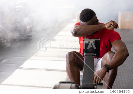 African American man wearing red t-shirt wiping sweat while rowing on rowing machine at gym African American man wearing red t-shirt wiping sweat while rowing on rowing machine at gym 140564305