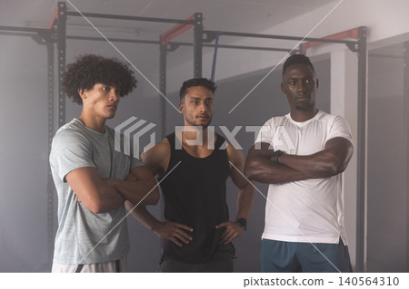 Diverse male friends standing in gym wearing smartwatch and athletic gear near steel power racks Diverse male friends standing in gym wearing smartwatch and athletic gear near steel power racks 140564310