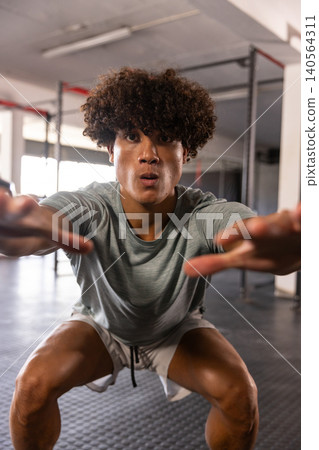 African American man squatting inside gym weight area wearing grey sportswear near metal power rack 140564311