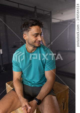 Asian male athlete sitting on plyometric box in gym showing smartwatch, squat racks, weight bench Asian male athlete sitting on plyometric box in gym showing smartwatch, squat racks, weight bench 140564314