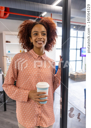 African american woman leaning against glass door in office by white shelving with coffee cup African american woman leaning against glass door in office by white shelving with coffee cup 140564329