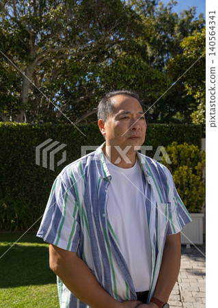 Asian man standing in sunlit backyard garden beside white planter box, wristwatch Asian man standing in sunlit backyard garden beside white planter box, wristwatch 140564341