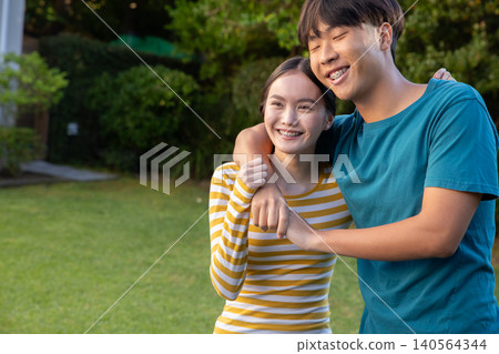 Asian couple standing on lawn in garden wearing casual clothing braces smiling toward camera Asian couple standing on lawn in garden wearing casual clothing braces smiling toward camera 140564344