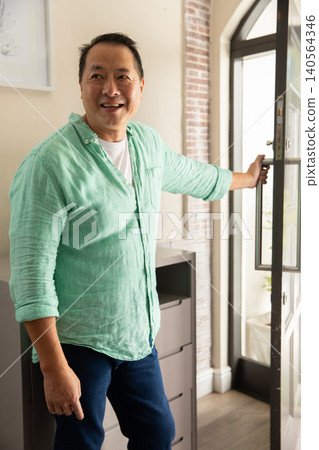 Asian man standing in home entryway holding glass-panel door beside chest of drawers with lamp Asian man standing in home entryway holding glass-panel door beside chest of drawers with lamp 140564346