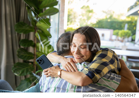 Diverse senior couple embracing on sofa by sliding glass door with smartphone, potted plant Diverse senior couple embracing on sofa by sliding glass door with smartphone, potted plant 140564347