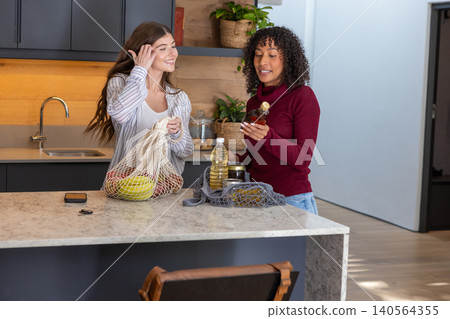 Diverse female friends unpacking mesh grocery bags at modern kitchen island with cooking oil bottle Diverse female friends unpacking mesh grocery bags at modern kitchen island with cooking oil bottle 140564355