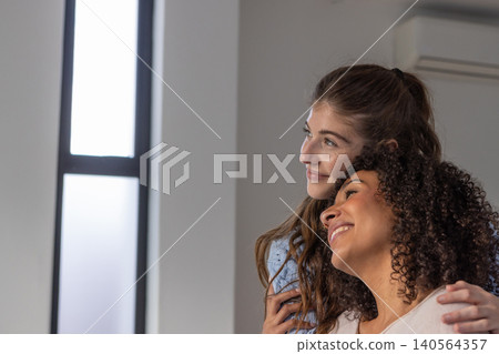 Diverse female friends leaning and gazing in living room with tall window and ac unit, copy space Diverse female friends leaning and gazing in living room with tall window and ac unit, copy space 140564357
