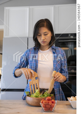 Asian woman mixing salad in wooden bowl using servers in kitchen with glass bowl of tomatoes Asian woman mixing salad in wooden bowl using servers in kitchen with glass bowl of tomatoes 140564367