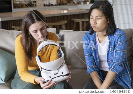 Diverse female friends examining white headset while sitting on beige couch in modern kitchen 140564368