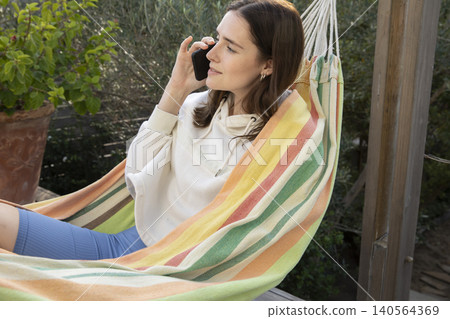 Woman in early 30s reclining on striped hammock on backyard deck talking on smartphone 140564369
