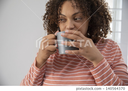 African American woman holding grey mug wearing striped top near blinds at table glancing sideways 140564370
