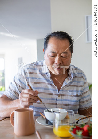 Mature asian man eating at table in bright kitchen using chopsticks with bowl, mug Mature asian man eating at table in bright kitchen using chopsticks with bowl, mug 140564371
