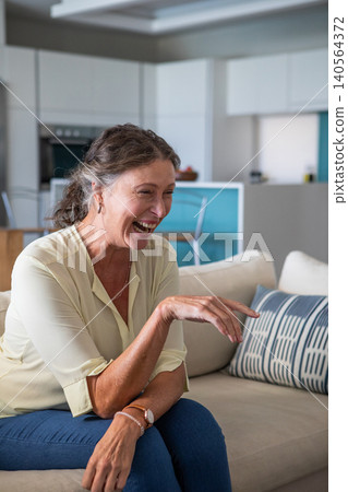 Senior woman laughing and pointing on cream sofa with pillow in kitchen with breakfast bar 140564372