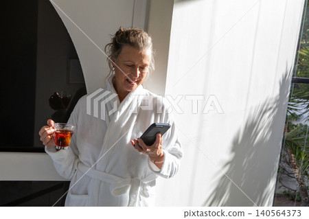 Senior woman standing in lounge holding smartphone, glass cup of tea by black screen Senior woman standing in lounge holding smartphone, glass cup of tea by black screen 140564373