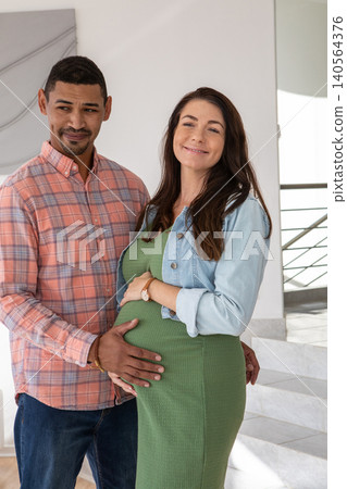 Diverse couple standing in modern home foyer touching baby bump by metal stair railing Diverse couple standing in modern home foyer touching baby bump by metal stair railing 140564376