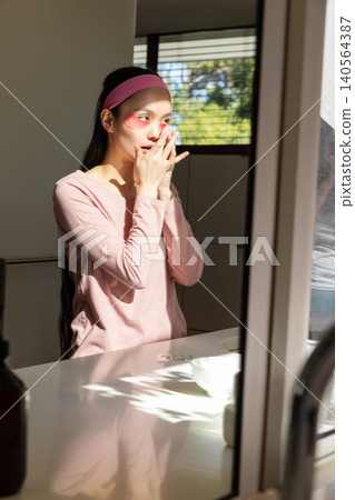 Asian woman wearing pink headband touching face with under-eye gel patches at vanity mirror Asian woman wearing pink headband touching face with under-eye gel patches at vanity mirror 140564387