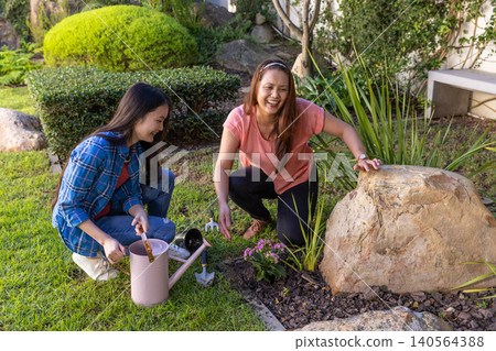 Asian mother and daughter planting flowering plant by garden boulder in backyard garden 140564388