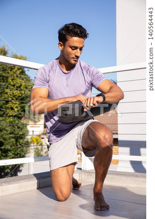 Fit man kneeling on rooftop balcony wearing lilac shirt holding yoga mat, checking smartwatch Fit man kneeling on rooftop balcony wearing lilac shirt holding yoga mat, checking smartwatch 140564443