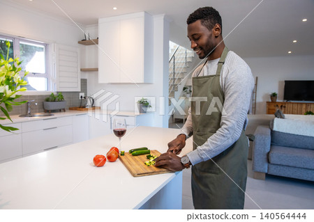 African american man slicing zucchini on board at kitchen island by tomatoes, wine glass African american man slicing zucchini on board at kitchen island by tomatoes, wine glass 140564444